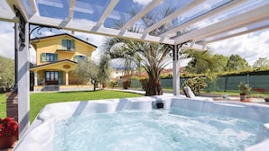 Un espace avec un jacuzzi, protégé par une paroi de verre, une maison jaune en arrière-plan et une pelouse bien entretenue.
