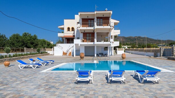A multi-story building with a swimming pool, blue lounge chairs, and a stone wall. - Villa Valasia in Gennadi (Gennadi)