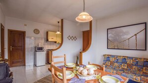 Zona pranzo con tavolo in legno apparecchiato per quattro persone, una cucina sullo sfondo e un divano colorato con cuscino decorato.