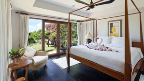 Un lit à baldaquin avec un ventilateur de plafond, une chaise en osier, une petite table sur laquelle est posé un vase de fleurs, et une vue sur un jardin par une porte vitrée.