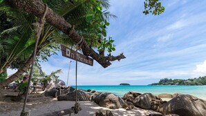 Une plage avec un panneau indiquant « Attention aux rochers », des palmiers et un ciel bleu dégagé.