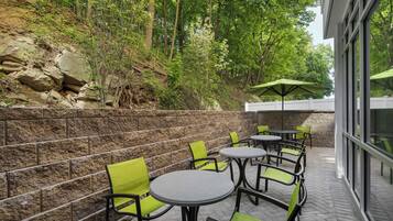 An outdoor patio with stone walls, green chairs, and tables, surrounded by trees.