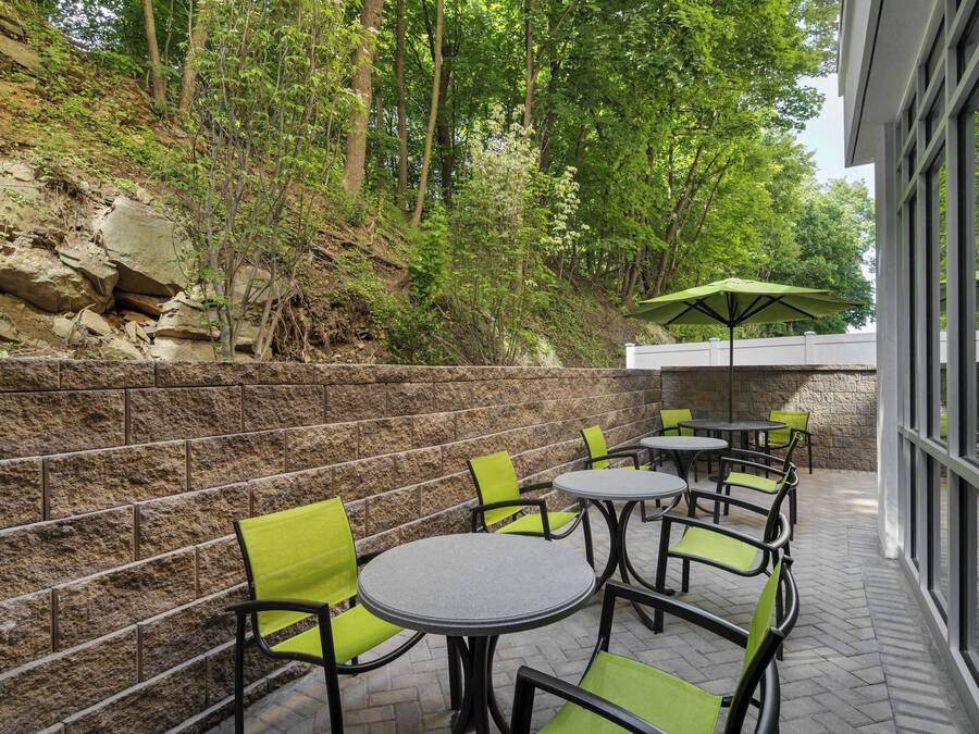 An outdoor patio with stone walls, green chairs, and tables, surrounded by trees.