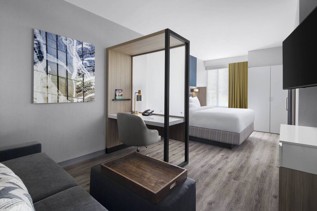 A modern hotel room with a bed, a desk, a chair, a coffee table, a TV, and a painting on the wall.