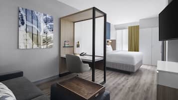 Una habitación de hotel moderna con una cama, un escritorio, una silla, una mesita de centro, un televisor y un cuadro en la pared.
