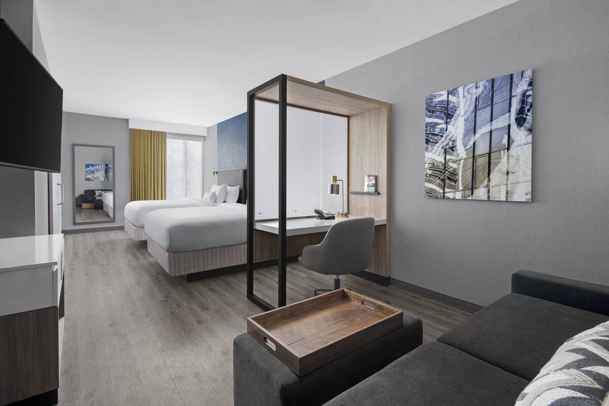 A modern hotel room with a large bed, a desk, a chair, and a small seating area.