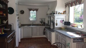 Una cocina con armarios blancos, piso de ladrillo, ventana con cortinas y diversos utensilios y electrodomésticos.
