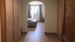 Un pasillo que conduce a un dormitorio con cama, ventana con cortinas y suelo de madera.
