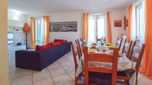 Uma sala de jantar com mesa posta para quatro pessoas, rodeada por cadeiras de madeira. Um sofá está posicionado junto à parede e um candeeiro é visível no canto.