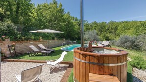 A wooden hot tub with a metal pole, surrounded by a brick wall and a pool. There are lounge chairs and a white umbrella in the background. - Il cacciatore di lucciole (Montecchio)