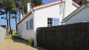 Un chemin de terre mène à un bâtiment blanc, doté de volets bleus et d’un toit recouvert de tuiles rouges.