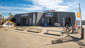 Un bâtiment sur lequel est apposée une enseigne indiquant « Cours – Location », avec une planche de surf appuyée contre.