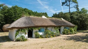 Une maison traditionnelle à toit de chaume, dotée d’un petit porche et d’une clôture en bois, entourée de verdure et d’arbres.