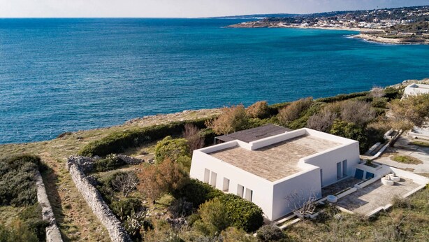 A modern white building with a flat roof, surrounded by greenery and a stone wall, overlooking a coastal area with clear blue waters and distant land. - Direct sea access and stunning location (Leuca)