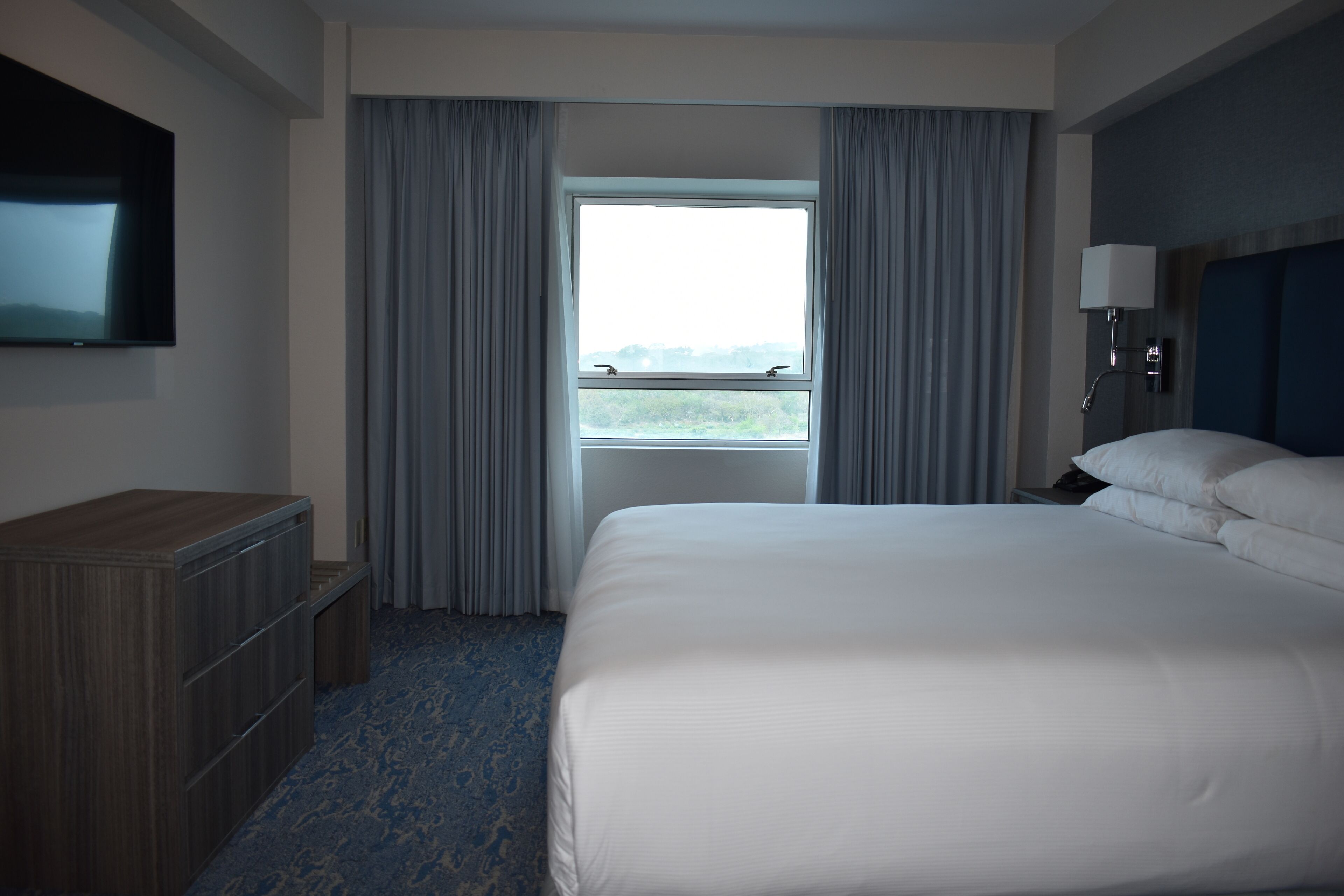 Habitación de hotel con una cama grande, un televisor montado en la pared, una ventana con cortinas, una mesita de noche con lámpara y un aparador de madera.
