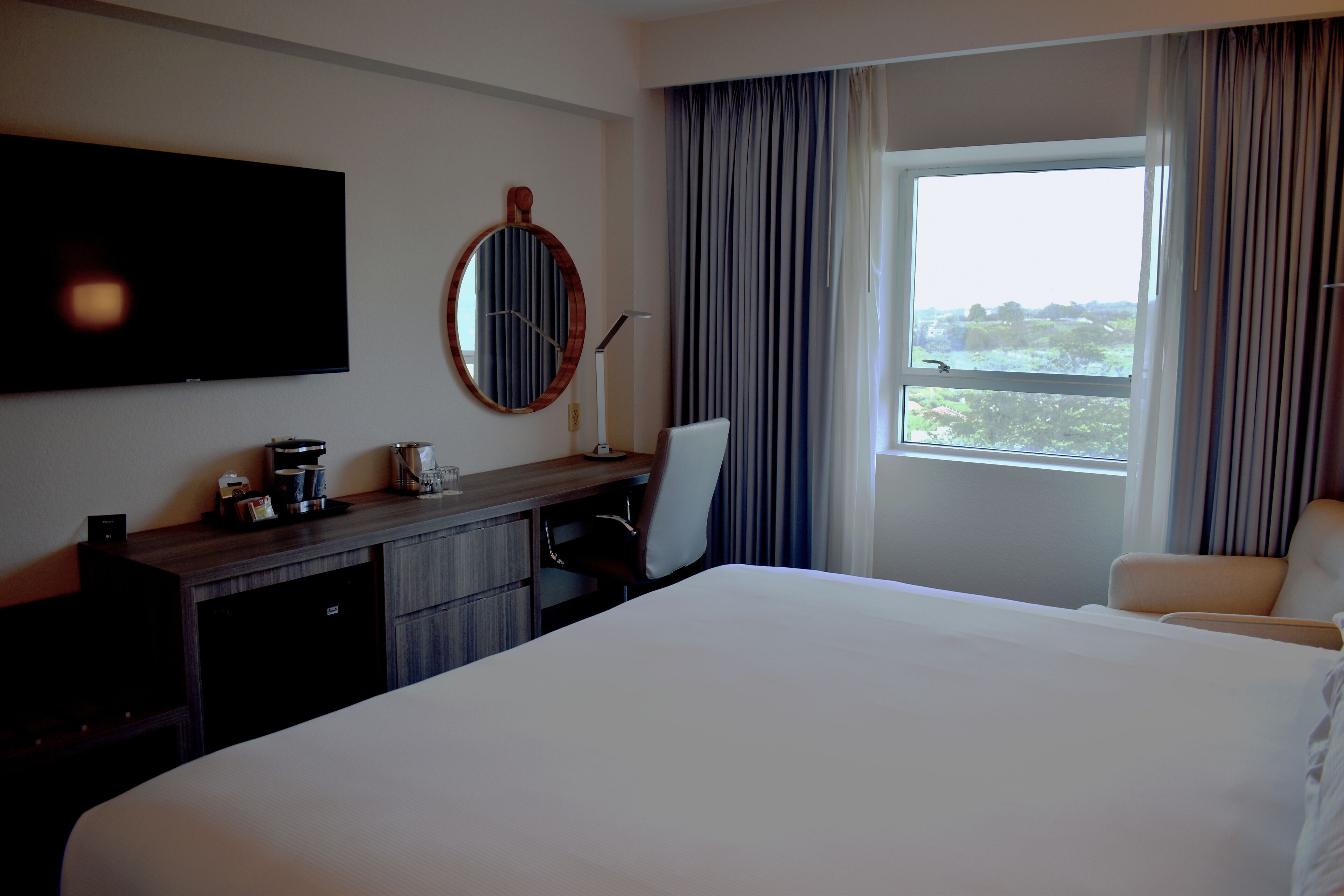 Una habitación de hotel con una cama grande, un escritorio con una silla, un televisor de pantalla plana, una cafetera y una ventana con cortinas.