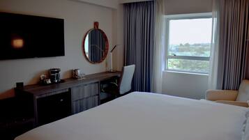 A hotel room with a large bed, a desk with a chair, a flat-screen TV, a coffee machine, and a window with curtains.