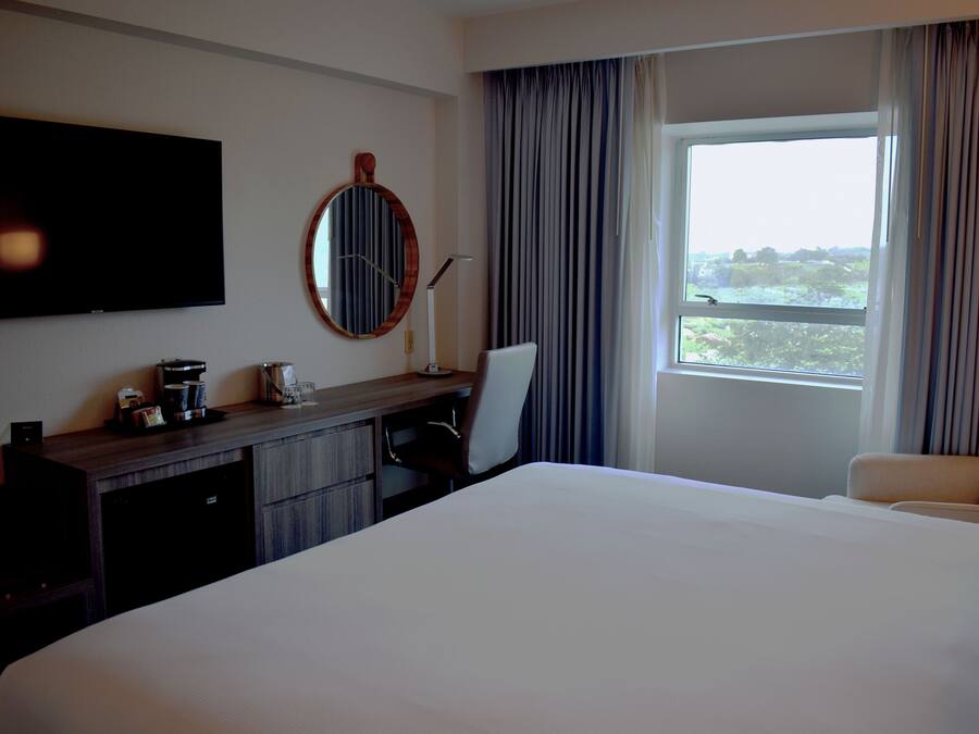 A hotel room with a large bed, a desk with a chair, a flat-screen TV, a coffee machine, and a window with curtains.