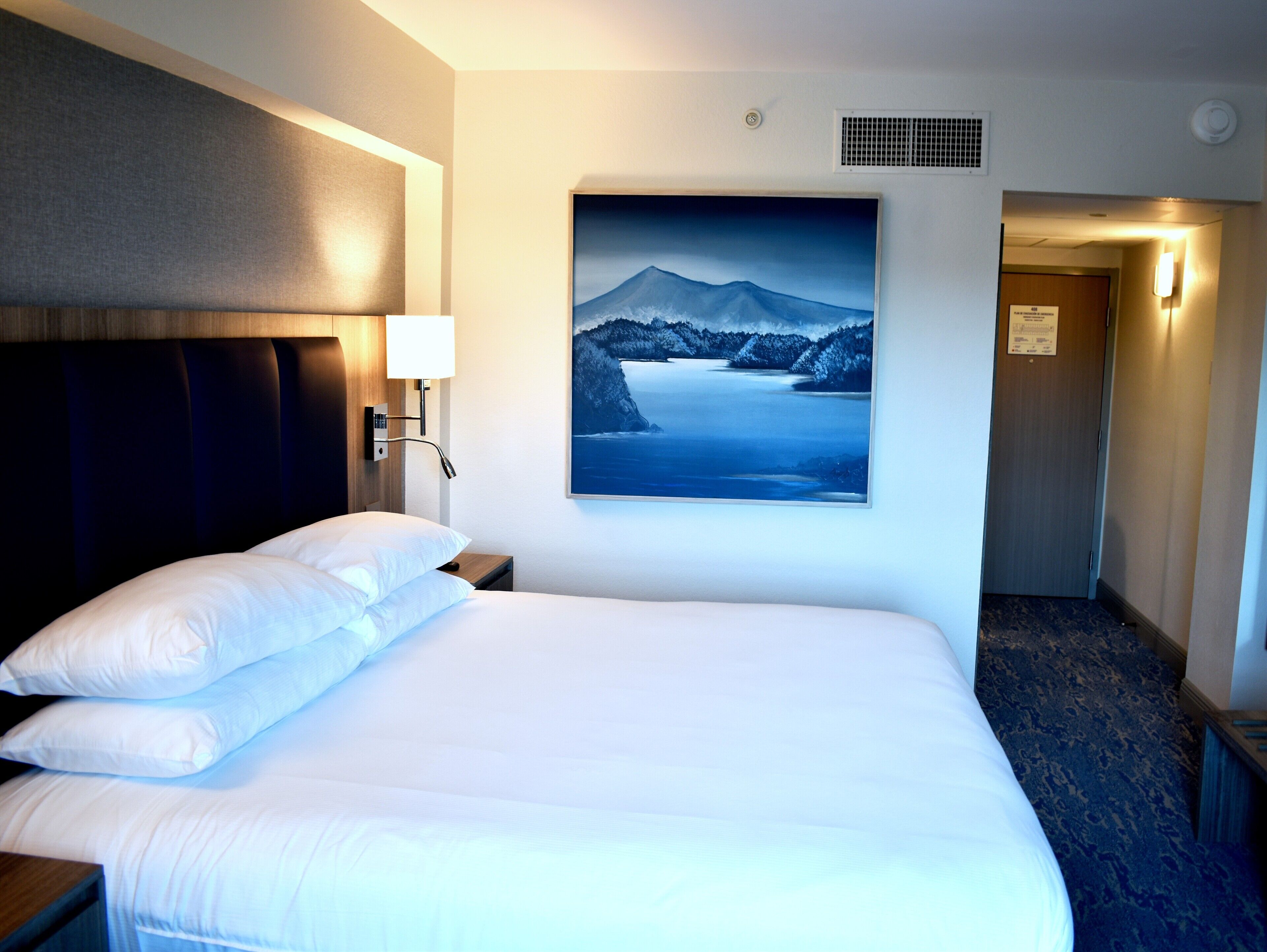 Ein Hotelzimmer mit einem großen Bett, einem Nachttisch, einer Wandlampe und einem Bilderrahmen mit einer Berglandschaft.
