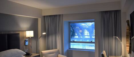 Habitación de hotel con cama, escritorio, silla, ventana con cortinas y lámpara.
