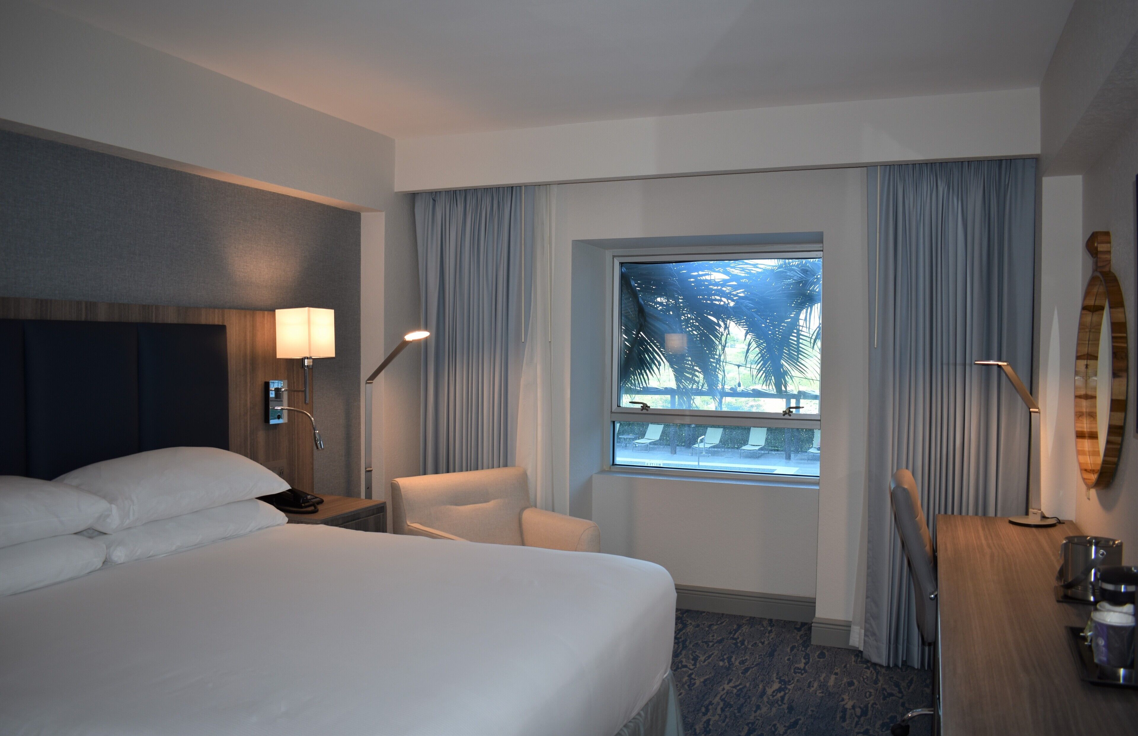 Ein Hotelzimmer mit einem großen Bett, einem Schreibtisch mit Lampe, einem Sessel und einem Fenster mit Vorhängen.