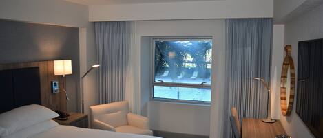 A hotel room with a bed, a chair, a desk, a lamp, a large window with curtains, and a view of the outdoors.
