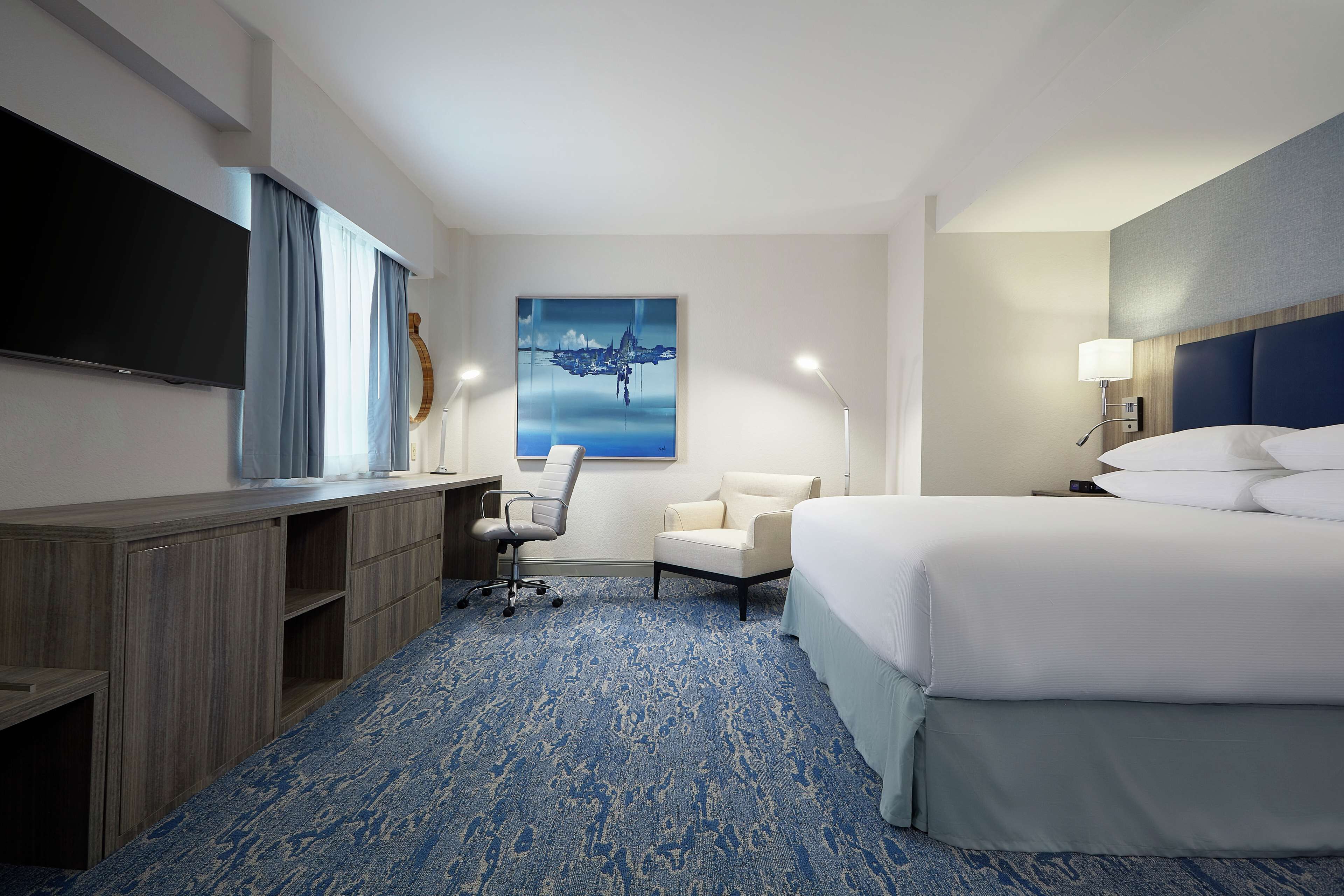 Une chambre d’hôtel avec un grand lit, un bureau avec une chaise, un téléviseur à écran plat, un tableau au mur et une lampe.