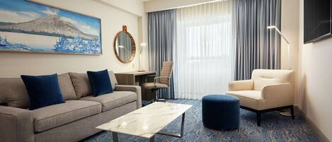 A hotel room with a sofa, armchair, ottoman, a small table, a desk, a chair, a TV, and a painting of a mountain.