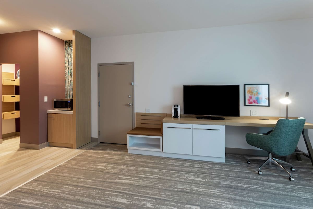 A modern hotel room with a wooden desk, a television, a green chair, and a wardrobe.