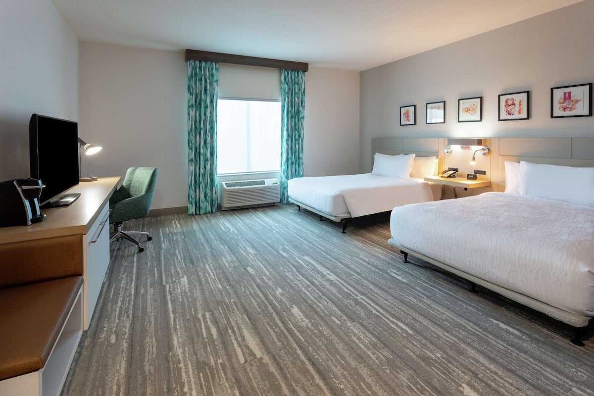 A hotel room with two beds, a desk with a computer, a chair, a window with curtains, and a wall with framed pictures.