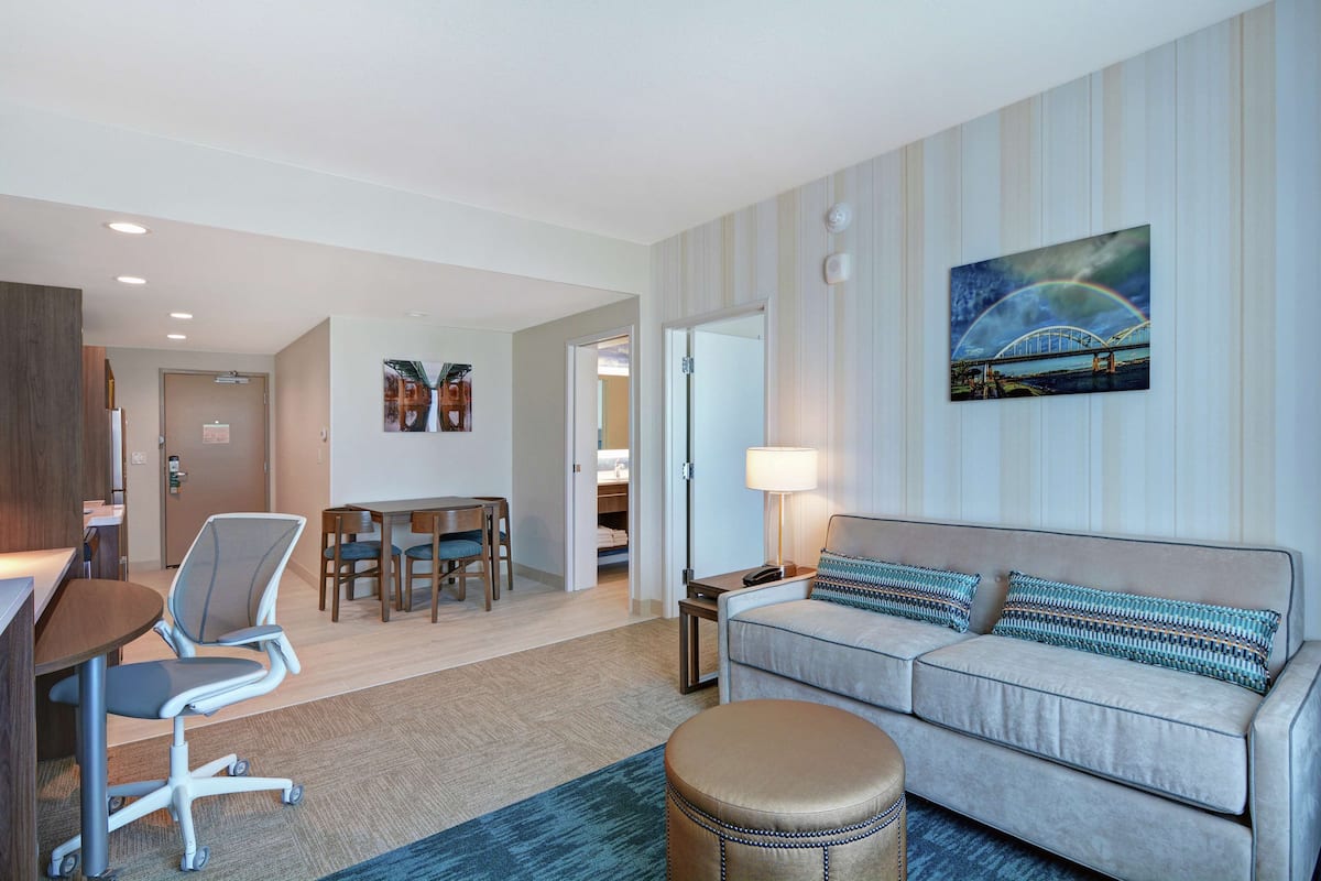 A hotel room with a sofa, a desk, a chair, a lamp, and a painting of a bridge.