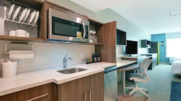 A modern kitchen with a microwave, sink, and coffee maker. There is a dining area with a chair and a bed in the background.