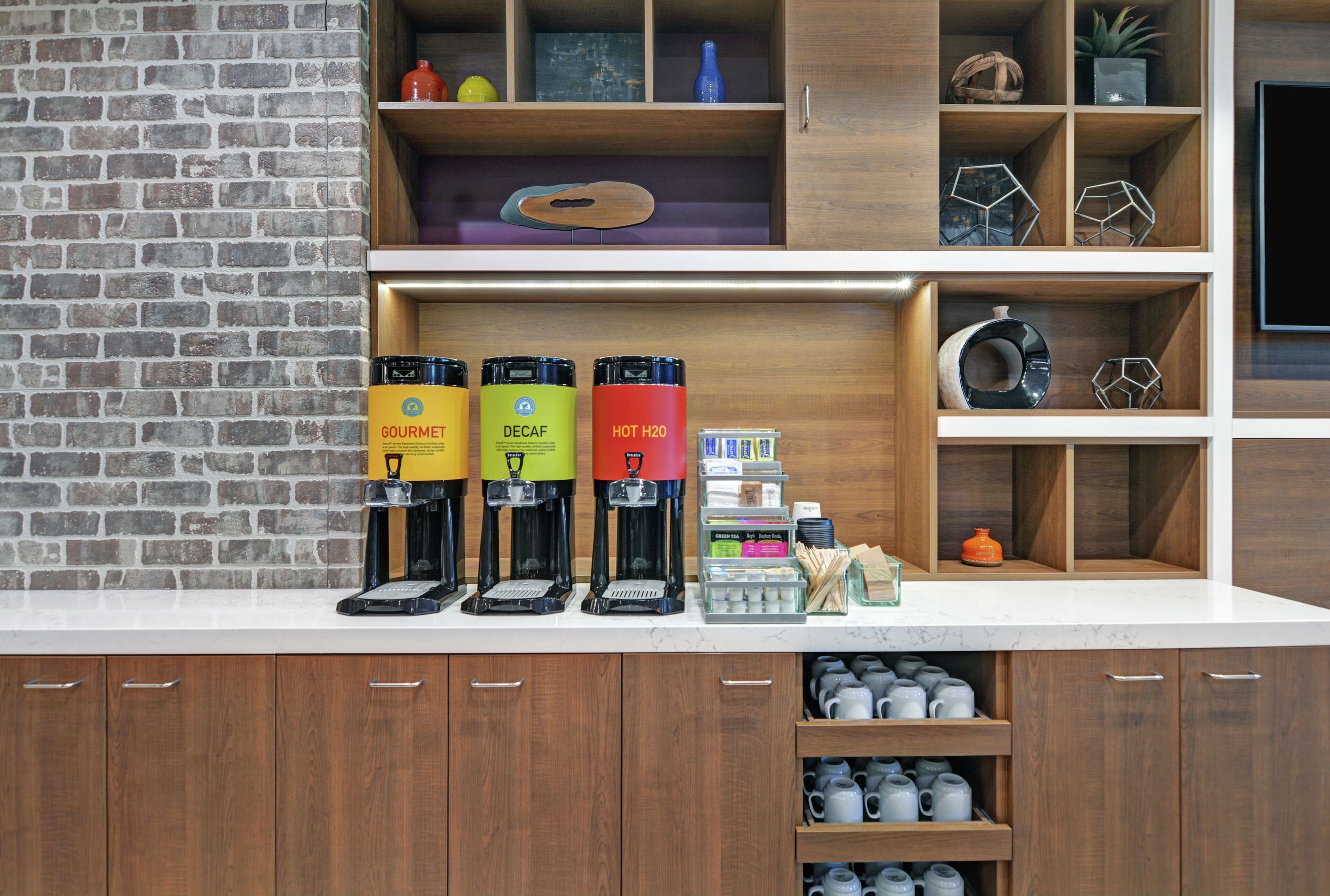 Een koffiehoek met drie koffieautomaten, gelabeld 'Gourmet', 'Decaf' en 'Warm H2O'. Er staan ook planken met diverse artikelen en een bakstenen muur op de achtergrond.