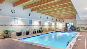 Une piscine intérieure avec des poutres en bois au plafond, des murs carrelés, ainsi que de nombreuses chaises et tables disposées le long du bord de la piscine.