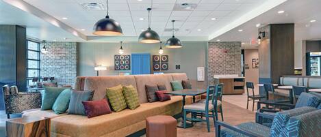 A modern lounge area with a large sofa, chairs, and tables, featuring a brick wall and pendant lighting.