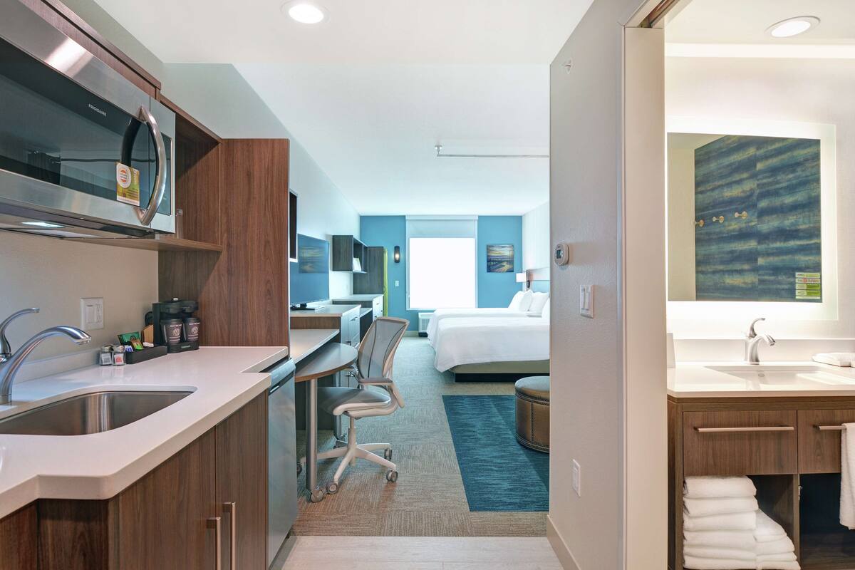 A modern hotel room with a kitchenette, a bed, a bathroom, and a view of the city.
