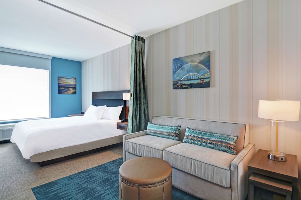 A hotel room with a large bed, a sofa, a small ottoman, a nightstand, and a lamp.