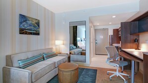 Une chambre d’hôtel avec un canapé, un pouf, un bureau et une chaise. Un tableau est accroché au mur et un miroir reflète la pièce.