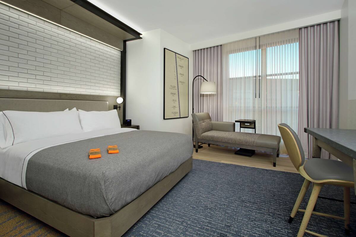 A hotel room with a large bed, a desk, a chair, a lamp, and a window with curtains.