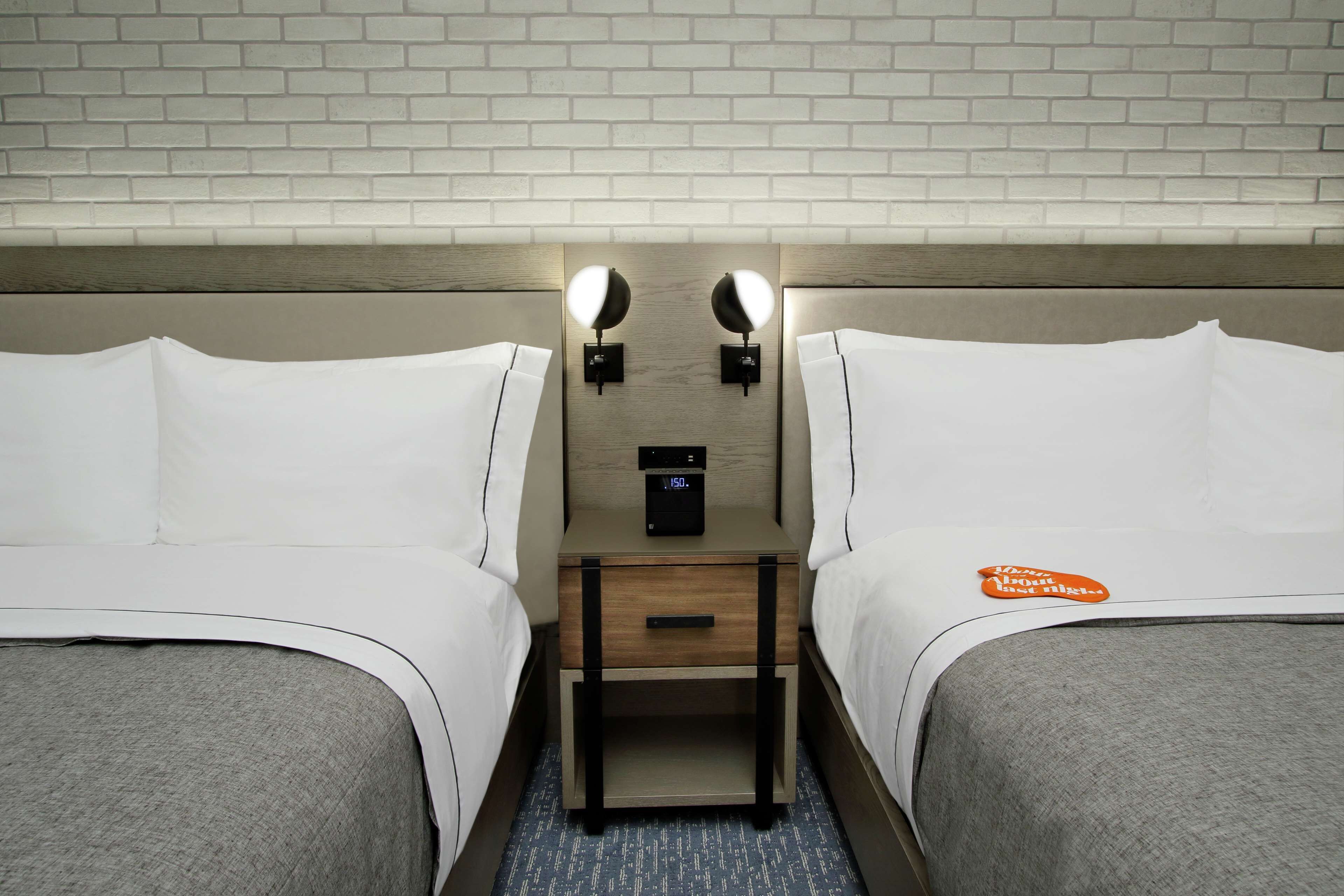 Habitación de hotel con dos camas, una mesita de noche de madera con un reloj digital y dos lámparas fijadas a la pared.