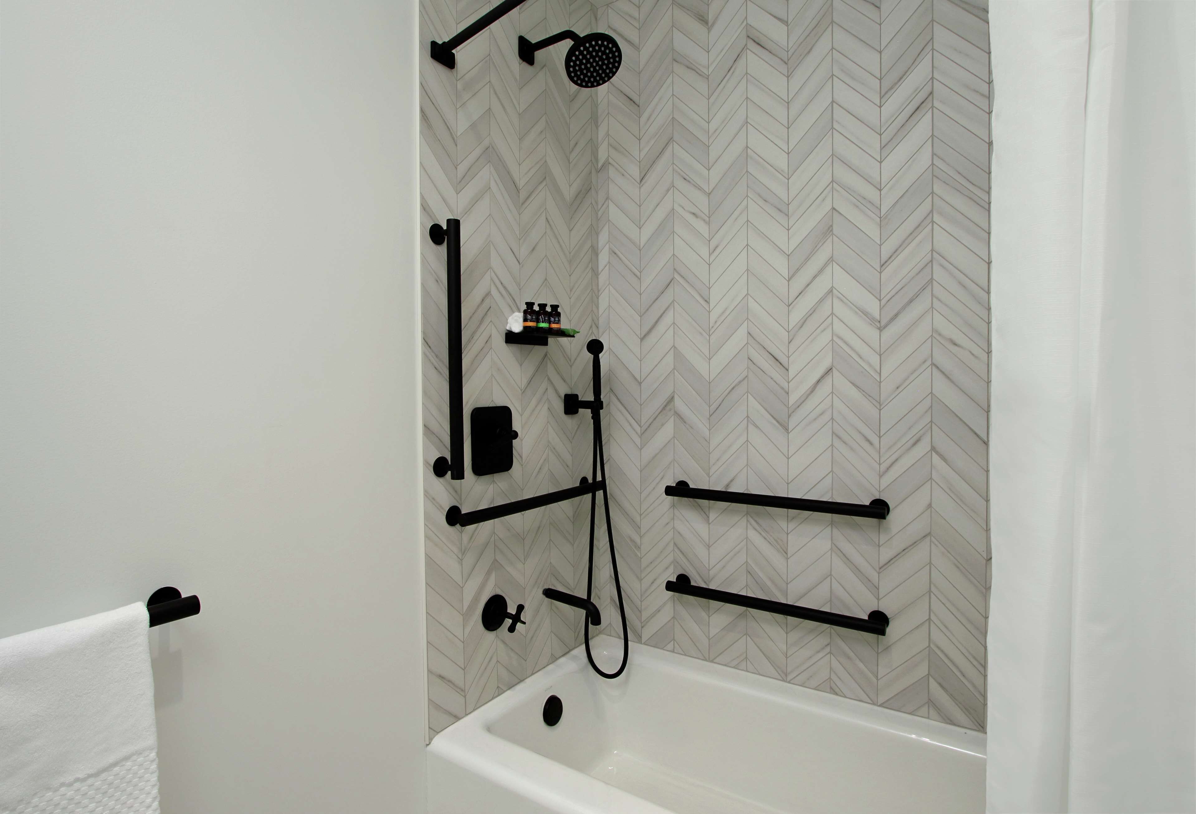 A bathroom with a white herringbone-patterned tile wall, a white bathtub, and black fixtures including a showerhead and towel racks.