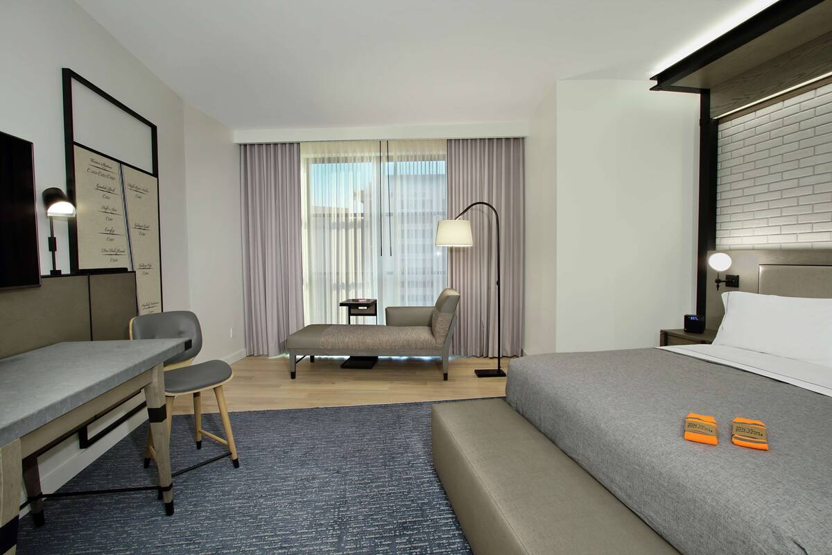 A hotel room with a bed, a desk, a chair, a small table, a lamp, and a window with curtains.