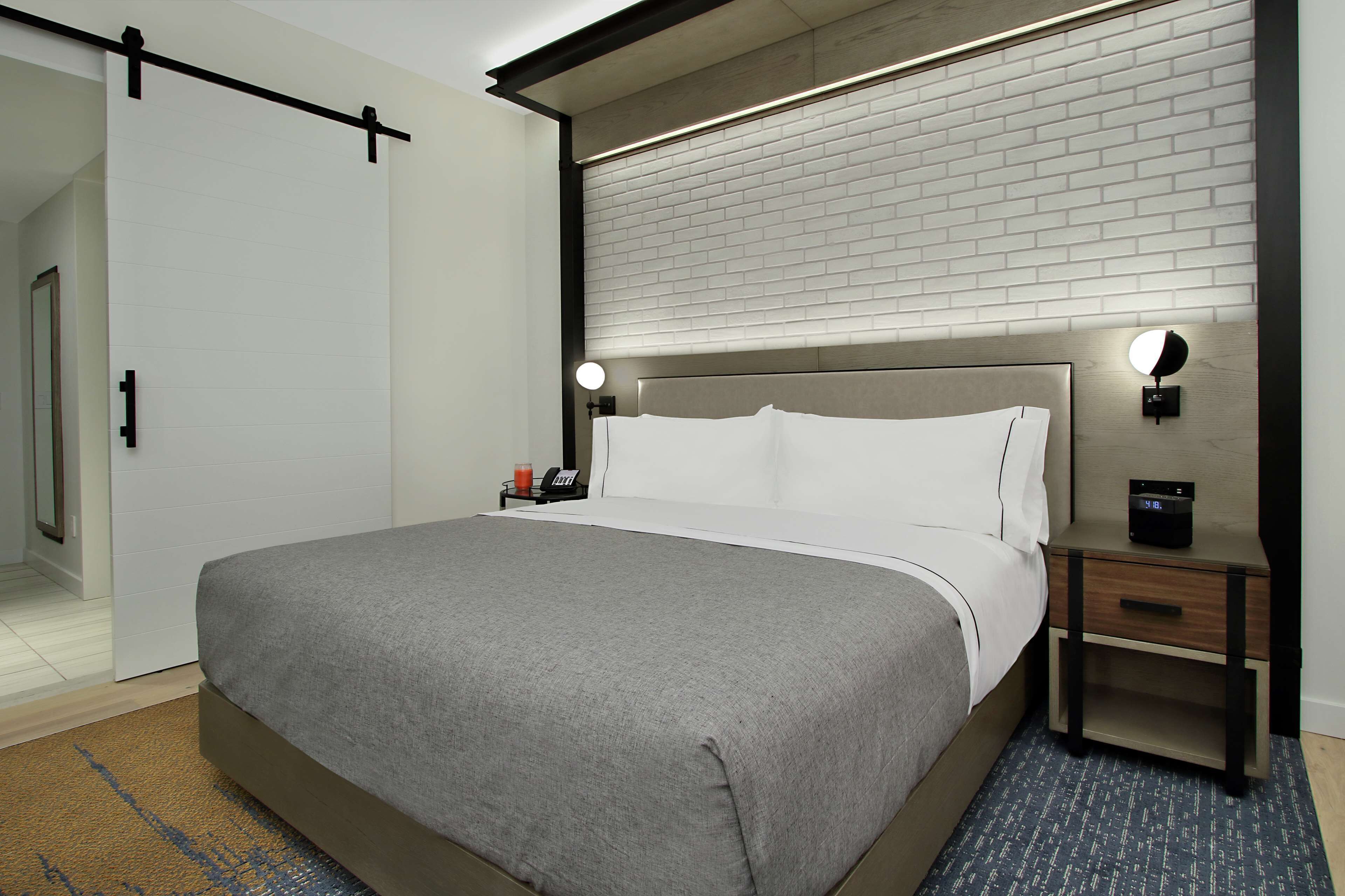 A modern hotel room with a large bed, a nightstand with a clock, and a wall-mounted light.
