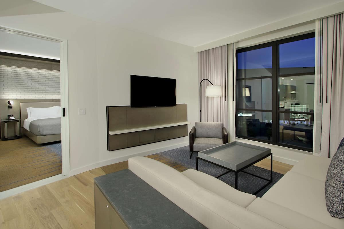 A modern hotel room with a sofa, armchair, flat-screen TV, bedside table, and a view of the city at night.