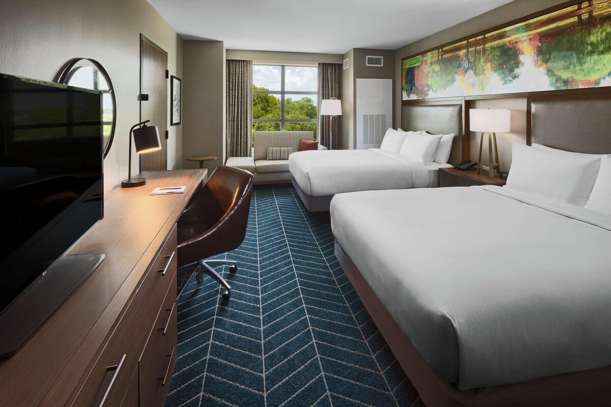 Ein Hotelzimmer mit zwei Betten, einem Schreibtisch mit Fernseher, einem Sessel und einem großen Fenster mit Blick auf Grünflächen.