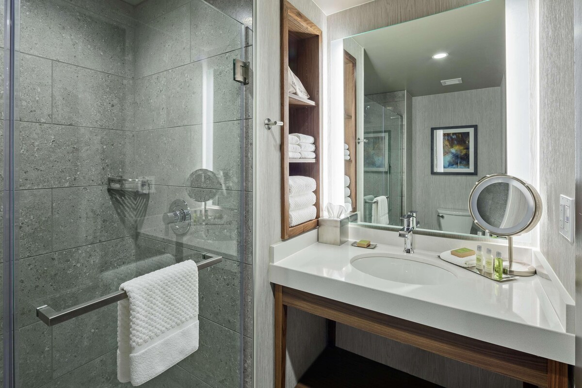 Une salle de bain moderne avec une douche à l’italienne en verre, un lavabo blanc avec un miroir et des serviettes soigneusement rangées.