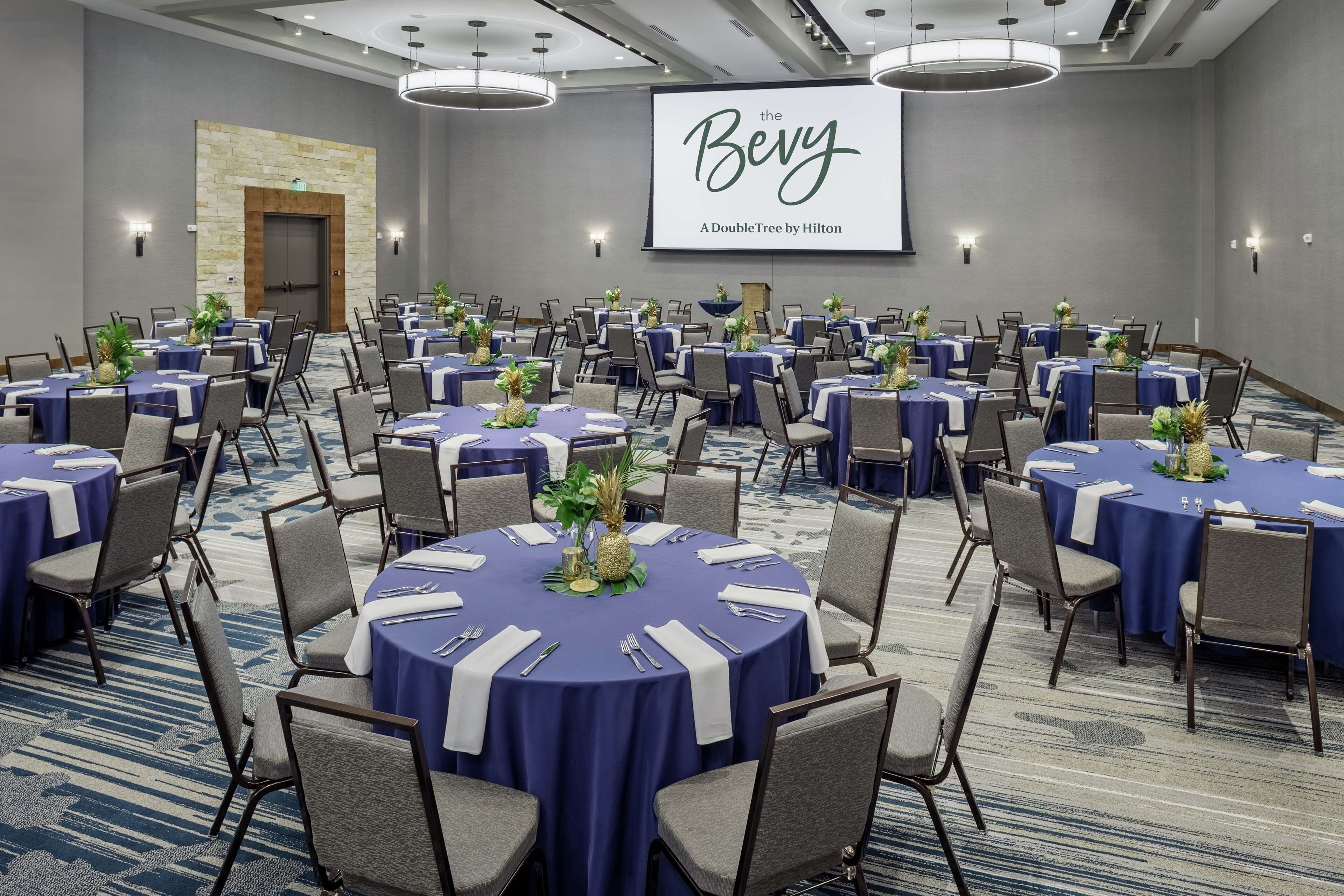 A conference room with round tables, chairs, and a large screen displaying "The Bery A DoubleTree by Hilton".