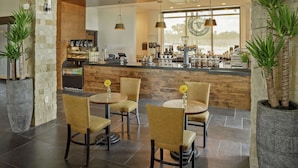 Un café moderne avec un mur en pierre, un plafond en bois et de grandes fenêtres. On y trouve des tables avec des chaises, un comptoir avec du matériel pour préparer le café et des plantes décoratives.