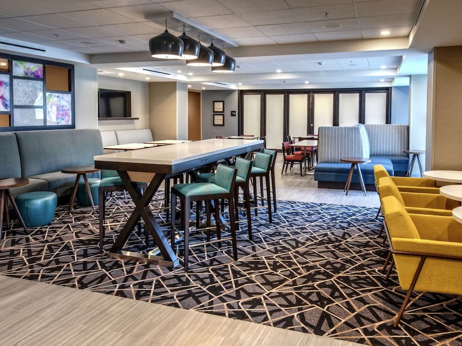 A modern hotel lobby with a long communal table, several seating areas, and a patterned carpet.
