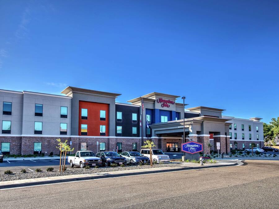 A Hampton Inn hotel with a modern design, featuring a mix of brick and glass facades, a parking lot with several cars, and a clear blue sky.